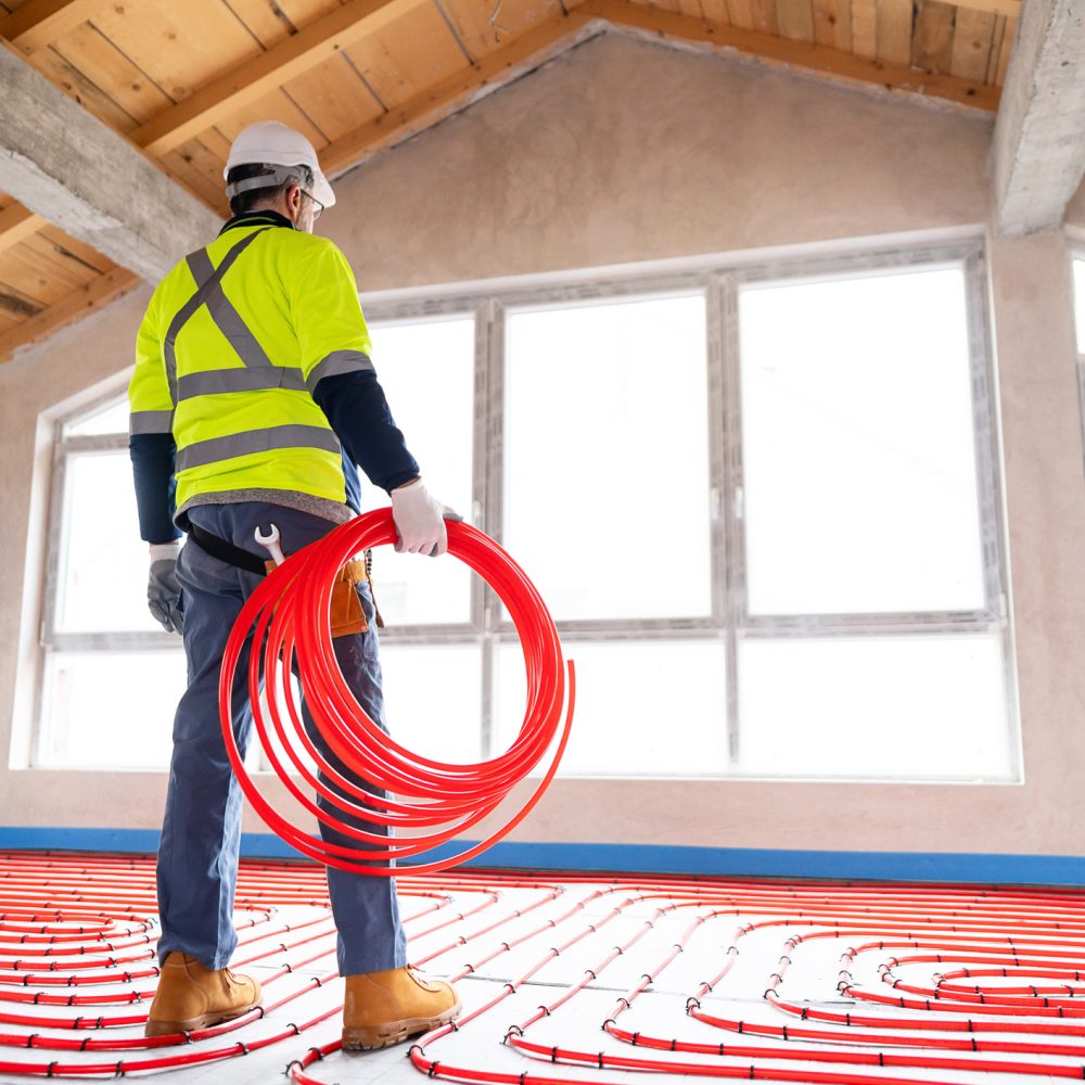 A construction worker in bright safety gear handles coiled red pipes for underfloor heating installation inside a mid-construction home with large windows and wooden ceiling.